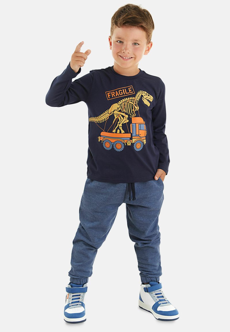 Blauses Langarmshirt mit einem Dinosaurier-Skelett-Grafik und dem Text "FRAGILE", kombiniert mit grauen Jogginghosen und weiß-blauen Sneaker-Stiefeln.