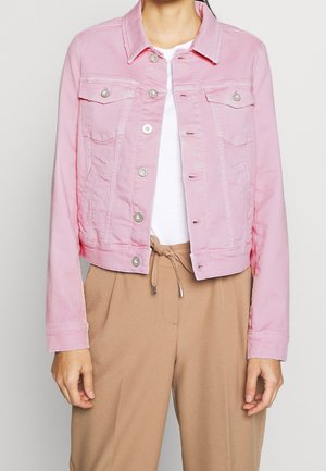 Personne portant une veste en jean rose courte sur une chemise blanche avec un pantalon beige à cordon de serrage, sur un fond uni.