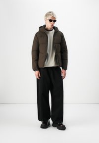 Brun pufferjakke med hætte, quiltet design. Bæret over en lys grå skjorte og sorte brede bukser. Accessories inkluderer sorte solbriller.