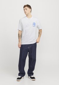 Camiseta de algodón blanca con estampado gráfico azul en el pecho, jeans de mezclilla oscuros de corte holgado y zapatillas negras con detalles blancos.