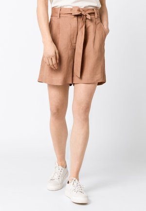 Femme portant un short marron taille haute avec une ceinture nouée et des baskets blanches, debout devant un fond blanc uni.