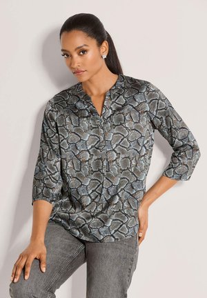 Vrouw met donker haar die een blauw-grijze blouse met slangenprint en driekwart mouwen en grijze spijkerbroek draagt, poserend tegen een lichte muur.