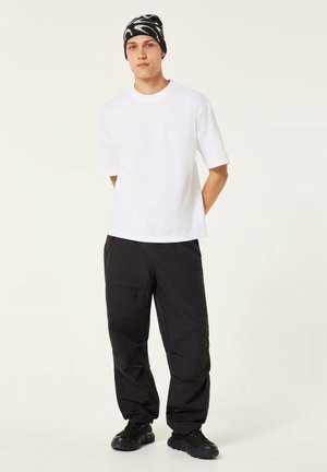 Un jeune homme porte une chemise blanche à manches courtes, un pantalon cargo noir, des baskets noires et un bonnet noir et blanc à motifs.