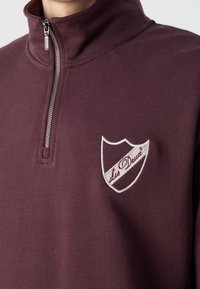 Sweat-shirt bordeaux à demi-zip avec fermeture éclair argentée et écusson logo blanc en forme de bouclier brodé "Les Deux" sur la poitrine gauche.
