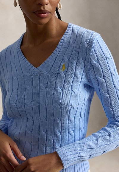 Žena nosi svetlomodrý sveter s výstrihom do V a žltým vyšívaným logom na hrudi, so škrupinovými náušnicami.