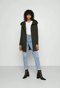 Mulher com cabelo encaracolado usa um casaco capuz verde escuro, uma blusa branca sem mangas, jeans em azul claro arregaçados e botas de tornozelo pretas de salto, de pé.