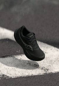 Mustad mustakate Saucony jooksujalats, mis on riputatud asfaldi kohal, valgete teedemarkidega ja väikeste killustikuga, mis lendavad all.