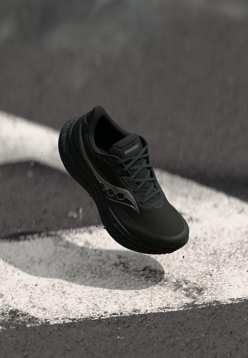 Mustad mustakate Saucony jooksujalats, mis on riputatud asfaldi kohal, valgete teedemarkidega ja väikeste killustikuga, mis lendavad all.