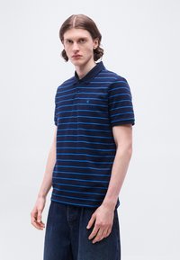 Νεαρός άνδρας με μεσαίου μήκους μαλλιά, φορώντας μπλε σκούρο πόλο με λεπτές ανοιχτό μπλε οριζόντιες ρίγες και σκούρο τζιν, στέκεται σε απλό φόντο.