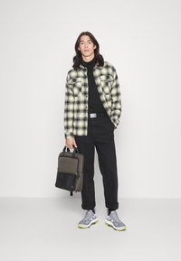 Gekacheltes Flanellhemd in Gelb und Schwarz, schwarzer Rollkragenpullover, schwarze Hose, grau-gelbe Sneakers und eine zweifarbige Umhängetasche.