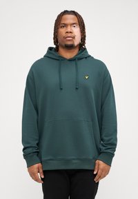 Sudadera verde de tejido suave con un bolsillo frontal, capucha con cordón y un pequeño logo amarillo en el pecho. Textura suave, corte relajado.