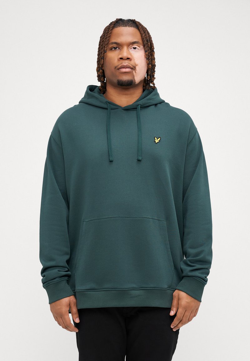 Sudadera verde de tejido suave con un bolsillo frontal, capucha con cordón y un pequeño logo amarillo en el pecho. Textura suave, corte relajado.
