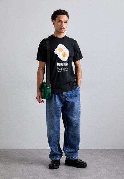 Black t-shirt featuring an egg graphic, paired with loose blue jeans. A green crossbody bag and black chunky shoes complete the outfit.