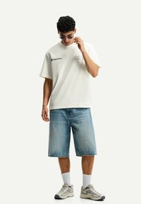 Creme t-shirt met korte mouwen en zwarte "Departures" tekst, gecombineerd met blauwe denim shorts. Model draagt grijze schoenen en een zonnebril.