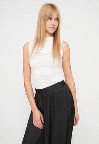 White sleeveless top with a high neckline, paired with black pleated wide-leg trousers. Smooth fabric with a subtle sheen and relaxed fit.