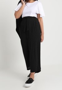 Pantalon large noir à pois blancs avec taille nouée, associé à un haut blanc à manches volantées et des chaussures noires sans lacets. Texture plate et lisse.