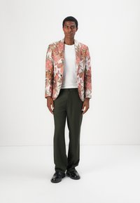Blazer mit floralem Muster in Pink und Grün, weißes T-Shirt, dunkelgrüne Hose und schwarze Schuhe. Der Blazer verfügt über einen Druckknopfverschluss.
