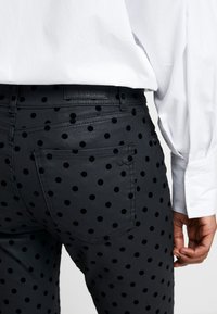 Persona vestida con pantalones negros con lunares negros y una camisa blanca de manga larga, mostrada desde la mitad de la espalda hasta la mano.