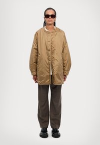 Beige quilted jacket with a soft sheen, featuring a button closure and two front pockets, paired with brown checkered pants and black chunky shoes.