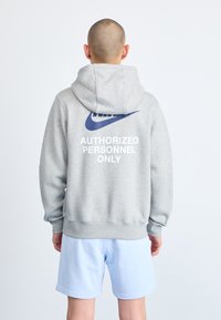 Siva bombažna kapuca z velikim modrim Nike emblemom in belim napisom "LE ZA POOBLAŠČENE OSEBE" na hrbtu. V kombinaciji s svetlo modrimi kratkimi hlačami.
