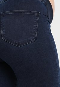 Jeans de mezclilla azul oscuro con un bolsillo trasero con un contorno cosido. El material tiene una textura suave con un desgaste mínimo.