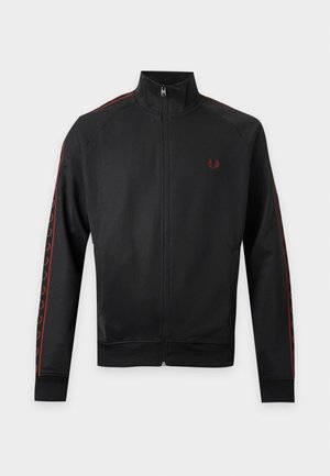 Joven con chándal negro con rayas rojas estampadas, gorra negra y zapatillas negras, de pie sobre fondo blanco liso.