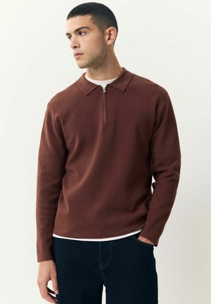 Suéter marrón de punto con cuello con cremallera, confeccionado en tela texturizada, combinado con una camiseta blanca y pantalones de mezclilla oscuros.