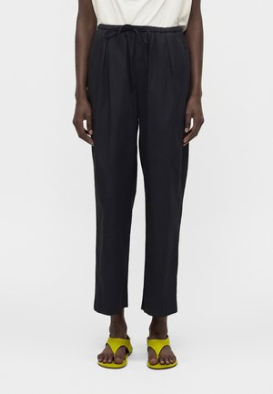 Άτομο που φοράει μαύρο ψηλόμεσο παντελόνι με δεμένη μέση και κίτρινες σανδάλες, στέκεται μπροστά σε λευκό φόντο.