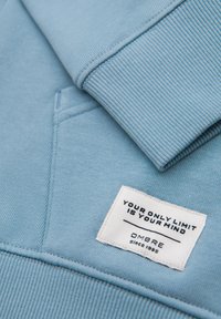 Felpa azzurra chiaro con colletto e polsini a costine, presenta un'etichetta con la scritta "IL TUO UNICO LIMITE È LA TUA MENTE" e il marchio "OMBRE dal 1985."