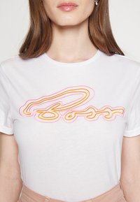 White short-sleeve T-shirt with a curved, multicolored logo design in pink and orange threads across the chest, featuring a smooth texture.