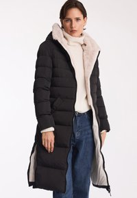 Padded, do kolan, czarny płaszcz z puszystym, jasnym kapturem. Posiada dwukierunkowy zamek błyskawiczny i boczne kieszenie. Nosi go na białym swetrze i dżinsach.