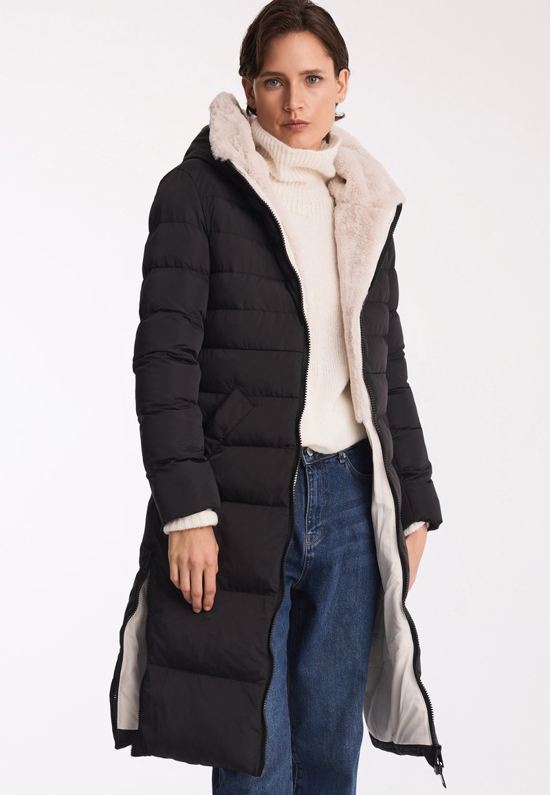 Padded, do kolan, czarny płaszcz z puszystym, jasnym kapturem. Posiada dwukierunkowy zamek błyskawiczny i boczne kieszenie. Nosi go na białym swetrze i dżinsach.