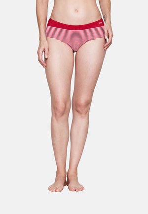 Frau trägt rot-weiß gestreifte Unterwäsche mit einem festen roten Taillenbund, steht barfuß vor einfachem Hintergrund.