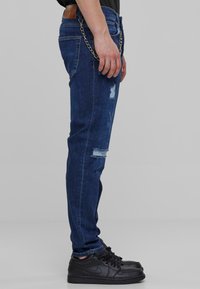 Mørkeblå denimjeans med slitte lapper, sølvkjedeaksent og standardlommer. Kombinert med svarte joggesko.