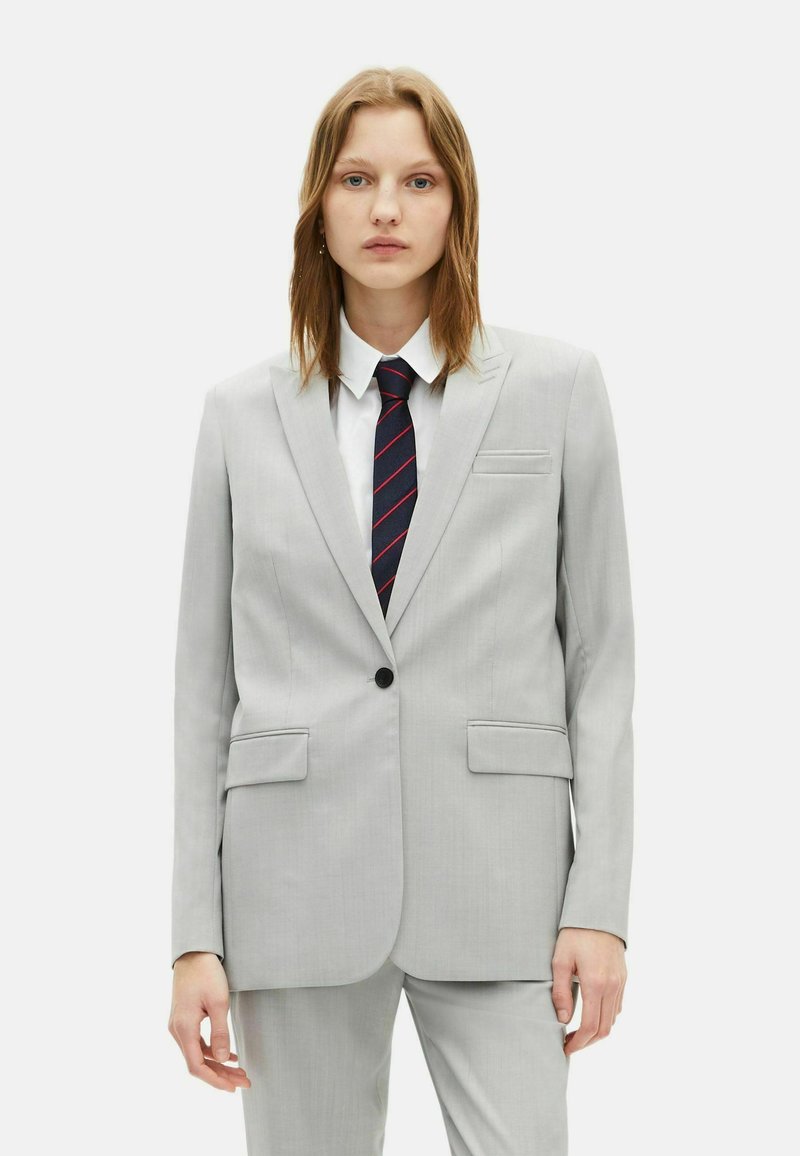 Completo grigio chiaro con blazer strutturato e pantaloni dritti; presenta due tasche frontali e una chiusura con un solo bottone; abbinato a una camicia bianca e una cravatta a righe.