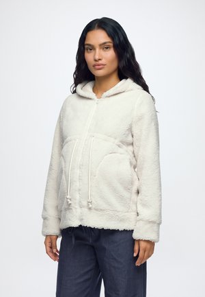 Mujer con cabello largo y oscuro que lleva una chaqueta de forro polar blanca con capucha y cremallera, con cordones y pantalones oscuros, de pie frente a un fondo liso.