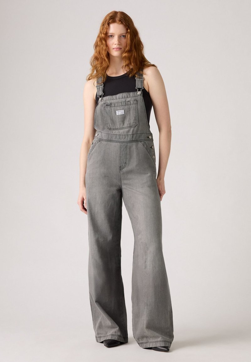 Grå denim overaller med vide ben, justerbare stropper, lomme foran og knapper på siden. Kombinert med en svart tettsittende tanktop.