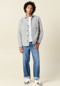 Veste en laine grise avec deux poches avant, fermeture boutonnée et col, portée avec un jean en denim bleu et des baskets blanches.