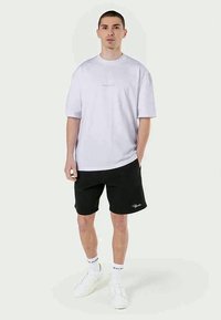 Vit t-shirt med kort ärm och avslappnad passform, matchad med svarta shorts med en vit logotyp, bärs med vita sneakers och ankelsockor.