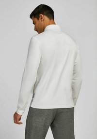 Weißes langärmliges Poloshirt mit weichem Material, klassischem Kragen und dezenten Seitenschlitzen. Darunter sind graue strukturierte Hosen zu sehen.