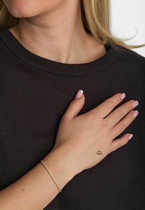 Črna majica s okroglim izrezom, na prsih je roka. Roka nosi nežno zlato zapestnico in ima vidno majhno tetovažo.