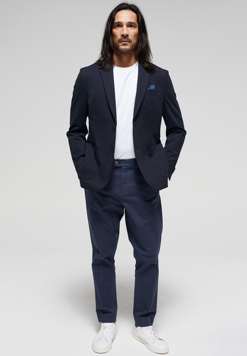 Hombre con cabello largo que lleva un blazer azul marino, camiseta blanca, pantalones azul marino y zapatillas blancas, de pie contra un fondo liso de color gris claro.