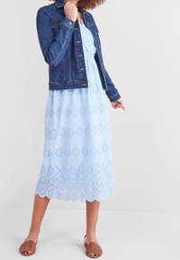 Jeansjacket over een lichtblauwe oogjesjurk met een golvende zoom, voorzien van opengewerkte ruitpatronen en bruine slidesandalen.