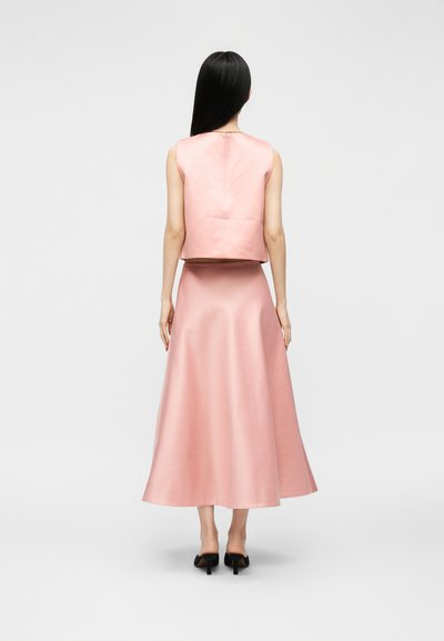 Γυναίκα με μακριά μαύρα μαλλιά, που φοράει ροζ αμάνικη μπλούζα και ροζ μίντι φούστα, στέκεται με πλάτη, φοράει μαύρα παπούτσια με τακούνι.