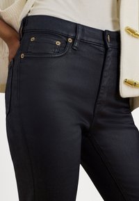 Tumši zili džinsa džinsi ar plānu piegriezumu, klasisku piecu kabatu dizainu, zelta toņa metāla detaļām un smalku tekstūru.