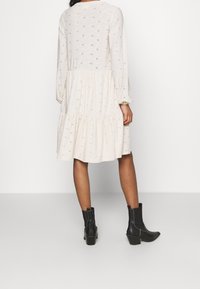 Robe crème à manches longues avec une jupe à volants, ornée de motifs brodés en forme de pois. Associée à des bottines noires.