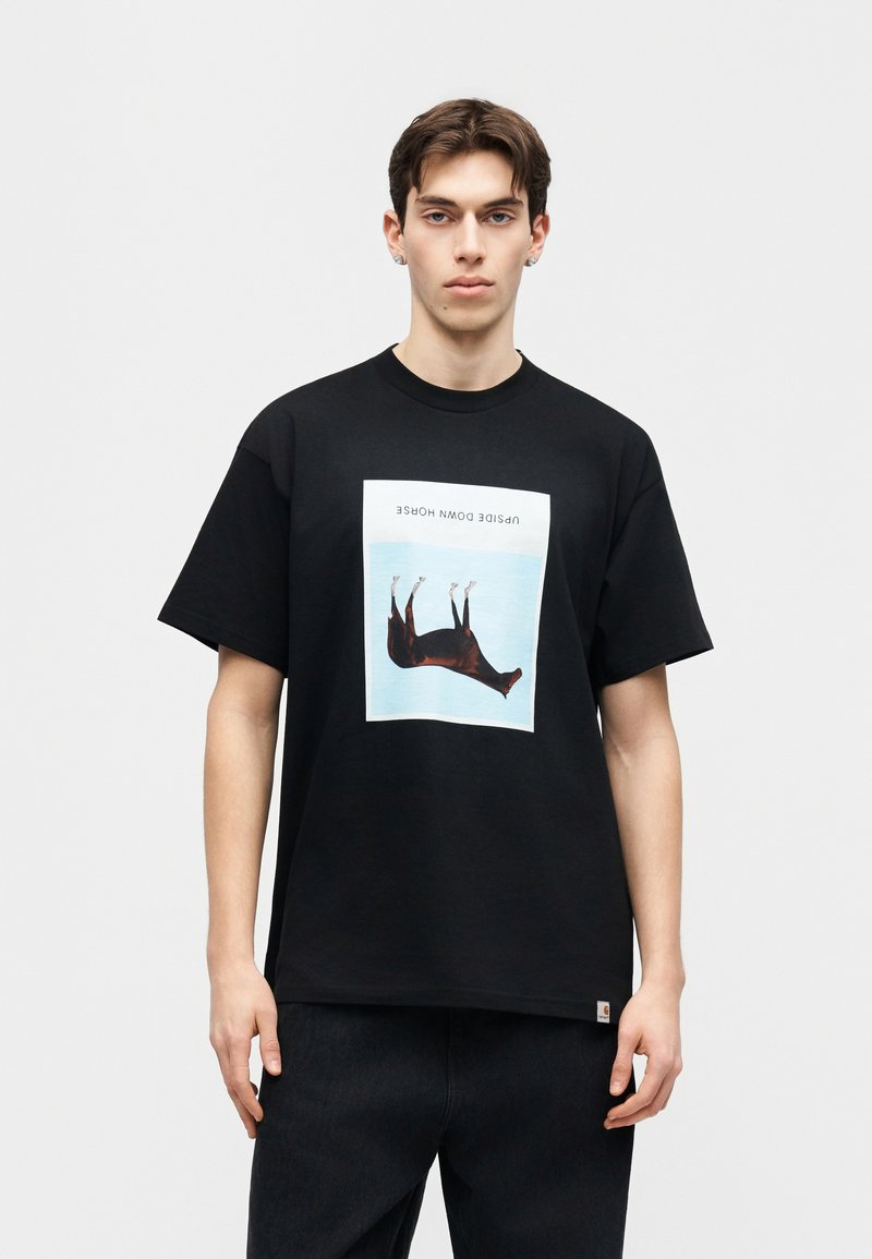 Junger Mann mit kurzen braunen Haaren, der ein schwarzes T-Shirt trägt, das eine Illustration eines auf dem Kopf stehenden Pferdes und den Text "UPSIDE DOWN HORSE" zeigt.