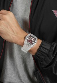Transparent watch with a square face, red and silver details, and a clear silicone strap, worn on a wrist against a black jacket and gray top.