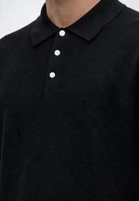 Polo en maille noire texturée avec trois boutons blancs et un col plat porté par une personne, cou et haut de la poitrine visibles.