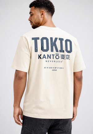 Mann trägt cremefarbenes T-Shirt mit "TOKIO KANTO" und geografischen Koordinaten auf dem Rücken, steht vor einfarbigem Hintergrund.